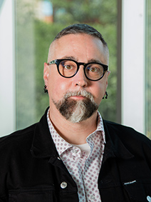 Portrait of a man with short-cropped black hair, a gray goatee, and black rimmed glasses wearing a black jacket and white and red dotted undershirt
