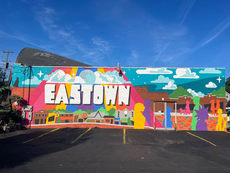 A colorful mural painted on the site of a building depicting a playful cityscape, colorful sillhouettes of people, and the word Eastwon in large white letters