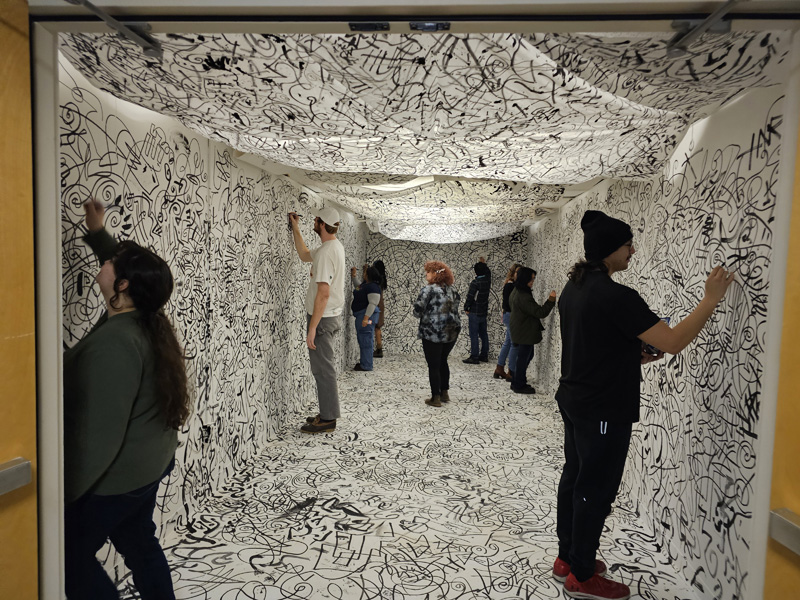 A group of people standing inside a white room in which the walls are covered with abstract doodles in black ink