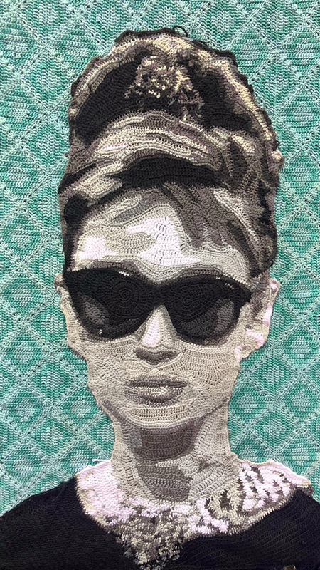 A portrait of actress Kathrine Hepburn, a woman with long brunette hair done up in a bun wearing a pearl necklace, sunglasses and black dress