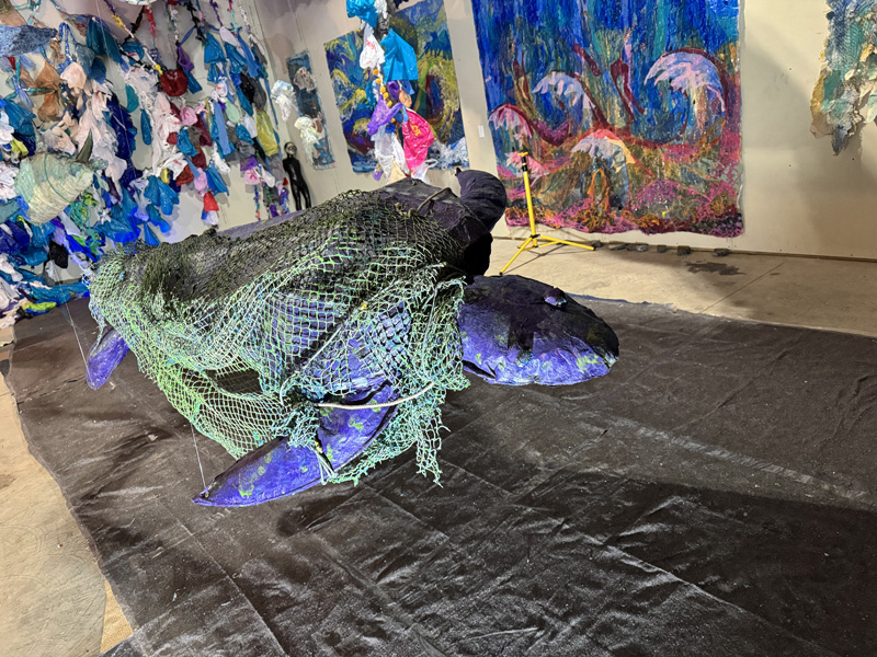 A sculpture of a sea turtle hanging from a ceiling, with colorful abstract paintings in the background