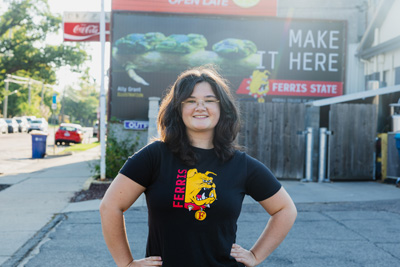 A young woman with black hair and glasses wearing a black t-shirt with a red and yellow bulldog on it standing in front of a billboard