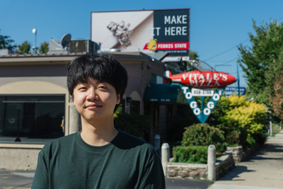 A young man with short black hair and a black t-shirt standing in front of a billboard