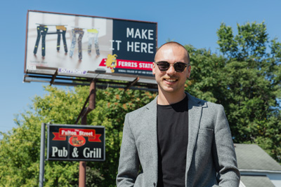 A young man with short brown hair and sunglasses wearing a gray blazer and a black t-shirt standing in front of a billboard