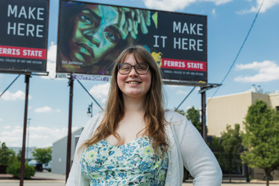 A young man with long brown hair and a black t-shirt standing in front of a billboard
