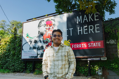 A young man with short black hair and a checkered-shirt standing in front of a billboard