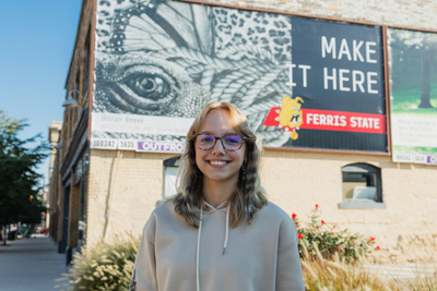 A young woman with long blonde hair black glasses and a cream sweatshirt standing in front of a billboard
