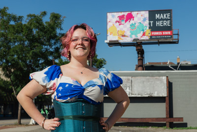 A young woman with dyed pink hair and a teal dress standing in front of a billboard