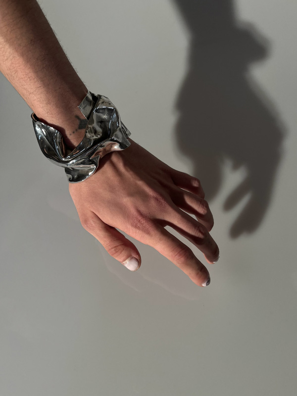 A photograph of a lower arm and hand wearing an elaborate, abstractly shaped silver bracelet. The hand casts a shadow on the white background.