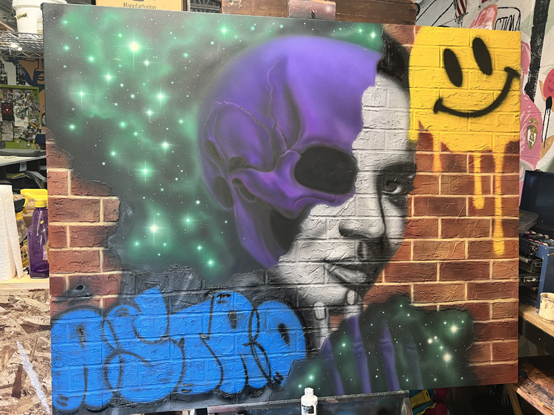 A spray-painted mural on a faux red brick wall depicting a smiley face, a female human face that morphs into a purple skull, elements of outer space, and the word astro rendered in purple and green graffitti style font 