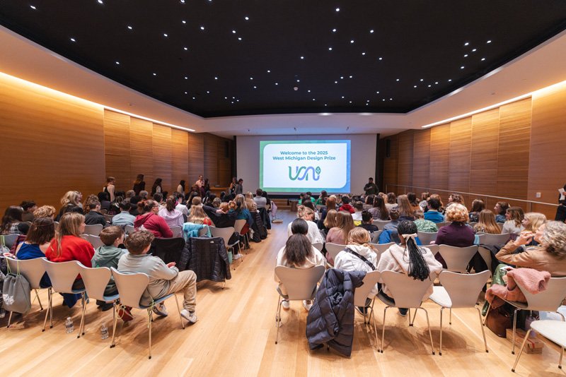 A large room with warm wooden walls filled with young students sitting in chairs staring attentively at a large screen on a stage