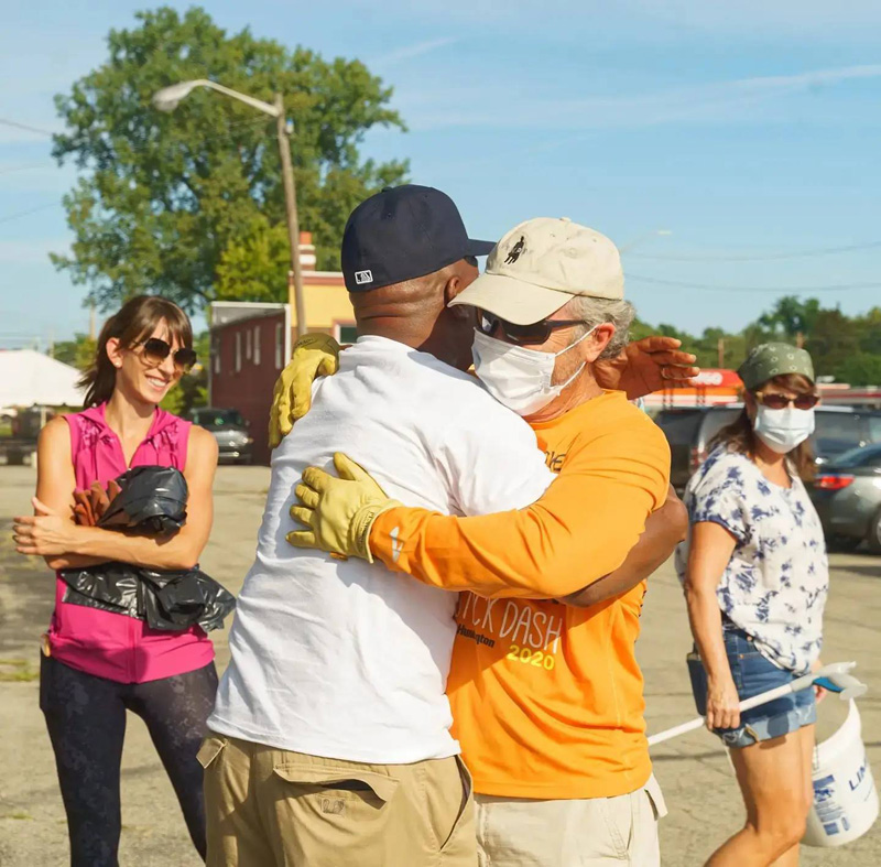 KCAD alum Abbey Chambers is shown participating in a community cleanup event through her social justice consulting firm, Justways Research. She stands to the side wearing a short sleeved pink top, black pants, and sunglasses while two men in the foreground embrace wearing work gloves and masks. 