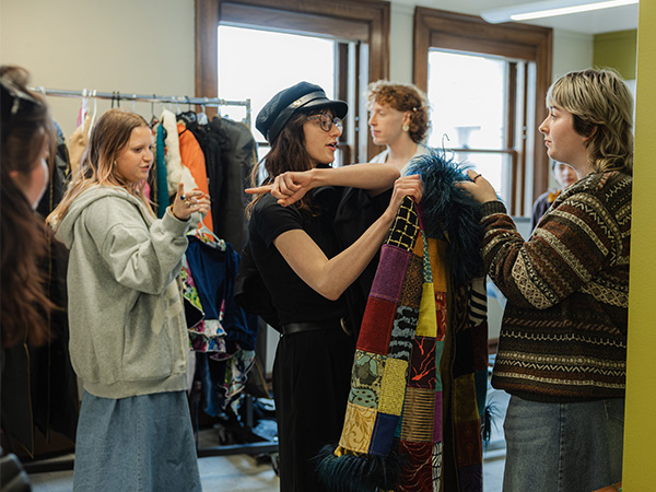 A group of students examining colorful garments in a clothing studio, with a person in a cap demonstrating a patchwork textile piece.