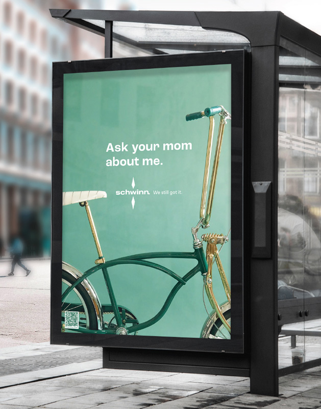 An advertisement situated on a city bus stop shelter that features a green background with an image of a vintage bicycle on it with the words ask your mom about me