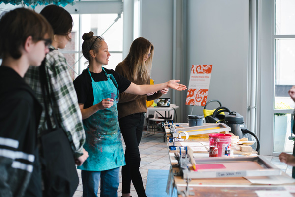 A diverse group of individuals gathered around a table, engaged in screenprinting activities led by a KCAD faculty member.