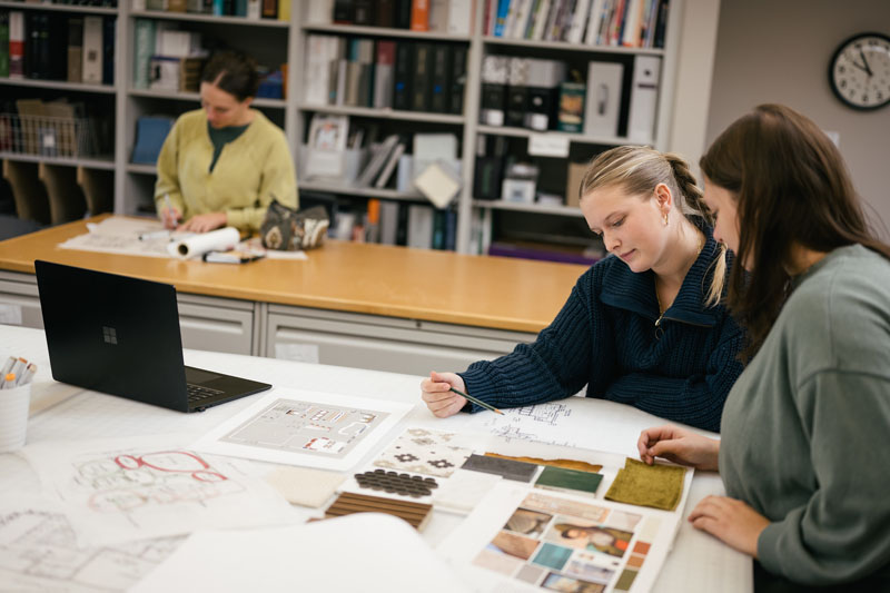 two young women, one with her long blond hair in a braided pony tail wearing a dark green zip up sweater and the other with shoulder-length brown hair wearing a light green sweatshirt, examine an interior design floor plan together 