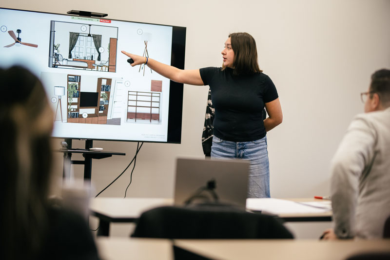 A young woman with shoulder length black hair wearing a black t-shirt and blue jeans gestures toward a large digital display showing an interior design floor plan