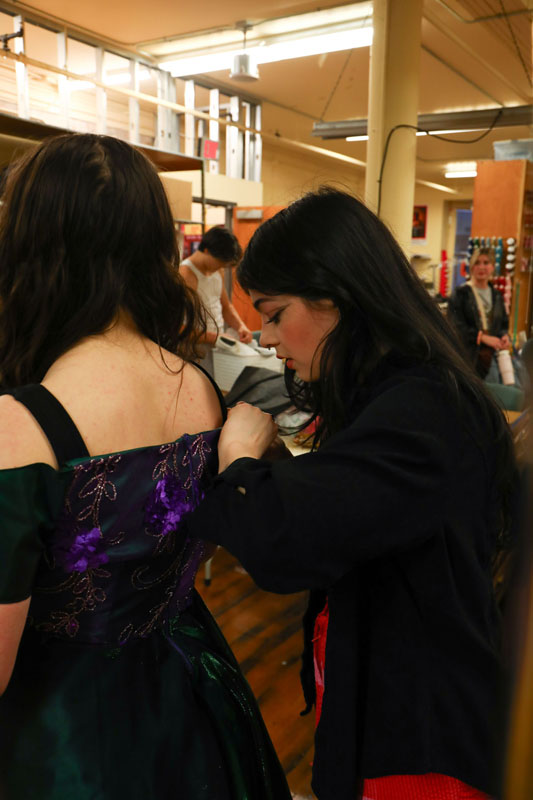 A young woman in a black sweater hunces over the back of an actor wearing a purple costume gown, pinning a booby pin