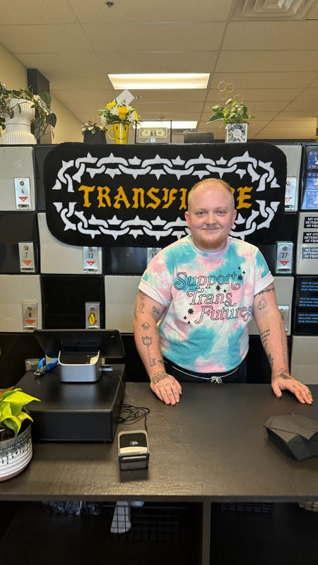 A trans man with close cropped hair and facial piercings standind behind a counter