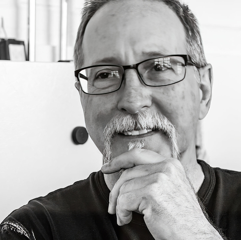 photograph of a man with short, spiky graying hear and a gray beard wearing a black t-shirt and black framed glasses