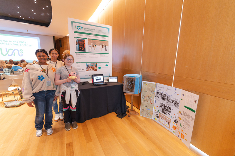 Three young women wearing white t-shirts pose next to an informational poster