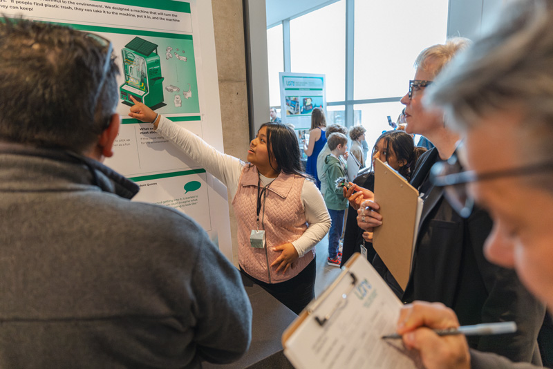 A young woman with long hair wearing a pink vest gestures toward an inforgraphic about a new recycling system as a crowd of adults looks on