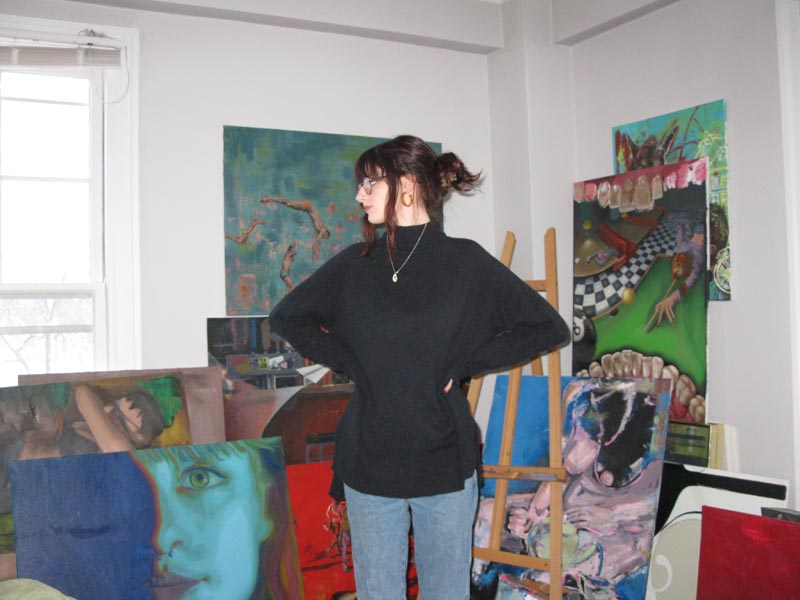 A woman wearing jeans, a black sweater, and glasses stands with her hands on her hips in a room filled with oil paintings