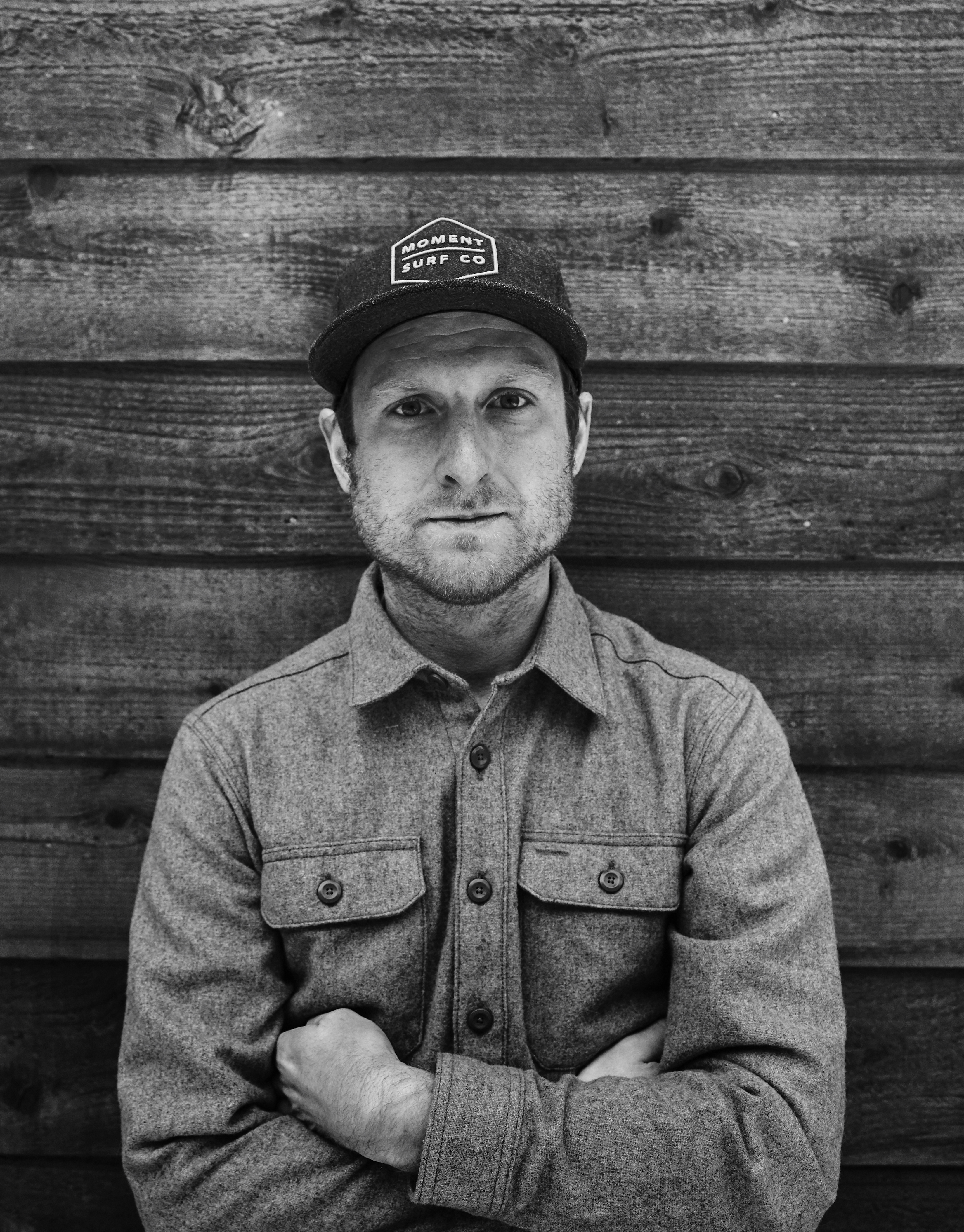 A portrait photograph of a man wearing a black baseball cap and gray long sleeve button up shirt. He has his arms folded and is looking solemly into the camera. 