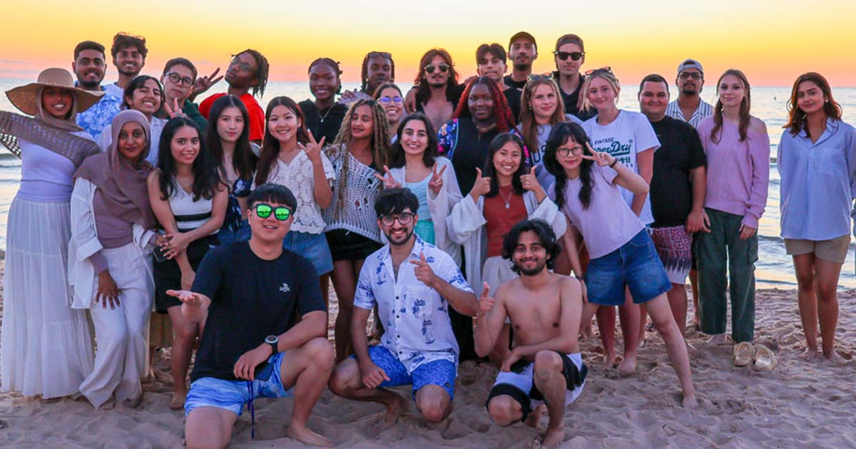 A group of Ferris State University international students standing in a group on a beach with the sun setting behind them in the background.